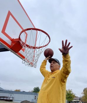 An older white man with a baseball hat, sunglasses, and yellow sweatshirt dunks a basketball.