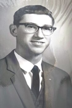 A black-and-white photo of Carroll as a young man with short black hair, rimmed glasses, a shy smile, and a suit and tie.