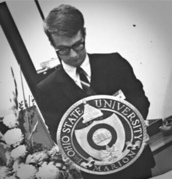 Carroll Neidhardt in a suit looks down at the logo he designed in cast metal. It has The Ohio State University Marion on the outer ring with a shield and Buckeye leaf inside.