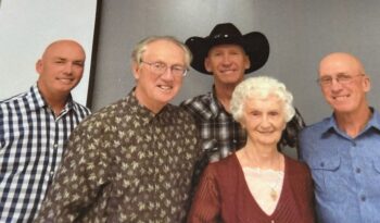 An elderly woman with soft white, curly hair smiles while surrounded by four taller sons.