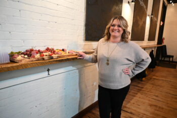 Leiha, a white woman with blond curly hair and a smile, a gray sweater, long gold necklace, and black pants, smiles at the camera while standing next to her charcuterie board loaded with meats, cheese, pickles, crackers, grapes and strawberries.