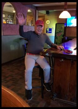 Dan is sitting on a bar stool with an OSU Hat, a big smile, a wave, a dark shirt and light jeans.