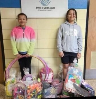 Two children smile while standing in front of the Boys & Girls Club Sign. In front of them are a large pile of Easter baskets.