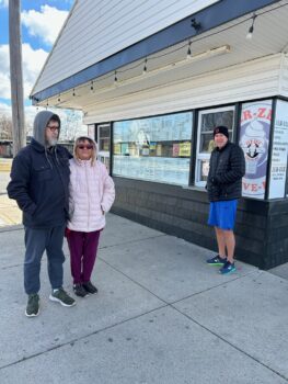 A couple and a single man stand in front of the Jer-zee. They are bundled up in coats. 