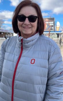 Kathy is a smiling white woman with short brown hair, sunglasses, and a gray puffy jacket with a small, red block "O".