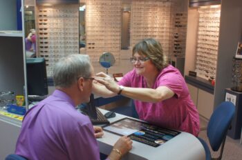 A female staff member smiles while helping a patient try on glasses. Rows of glasses are behind them. 
