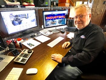 A white man with short white hair, a short white beard, and glasses, sits at a workstation with multiple monitors.