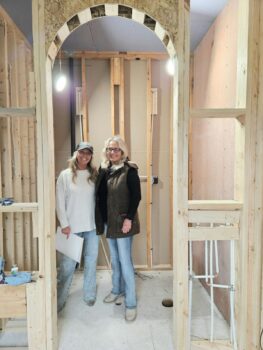 Two women stand and smile while in the middle of a home under construction. Blair is a white woman with a bright smile, ball cap, long blonde, wavy hair, a long white shirt, light demin jeans. Her mother is slightly taller with shoudler-length blonde hair, a black shirt, brown jacket, light blue jeans and gray boots.