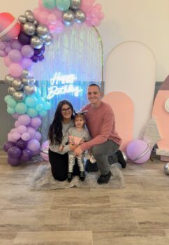 The family poses in front of a backdrop and balloon arch. Zach is a white man with a buz cut and smile. The daughter is a toddler with two brown pigtails and a smile. 