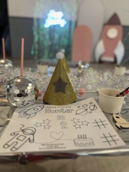 A table set for a toddler's birthday party with a coloring page, a bowl of crayons, a gold glittery party hat, a silver juice cup and more. 