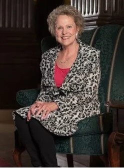 A white woman with short blonde curly hair, a wide smile, a pink shirt and black and white patterned jacket.