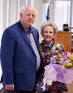 Pam smiles and holds flowers while standing next to her husband, a taller white man with short white hair, a gray polo shirt and a dark blue suitcoat. 