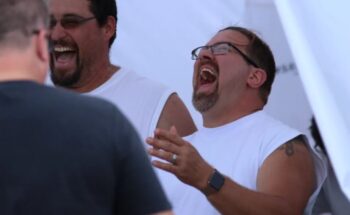 Two men laugh. Both have brown hair, goatees and glasses and white sleeveless shirts. 