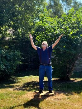 A man smiles with his hands raised while standing on a tree stump.