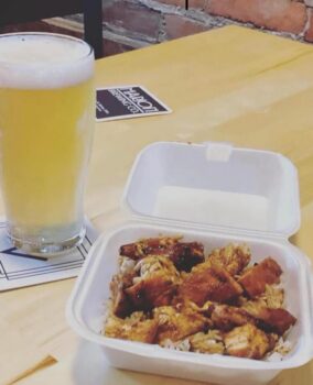 Photo of bourbon chicken on rice in a container next to a tall pint glass with a light-colored IPA.