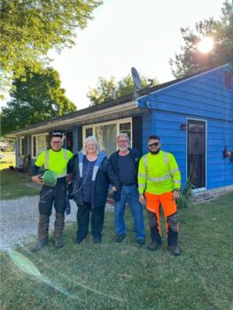 A woman with an oxygen tank and a man smile with the tree trimmers on either side of them. A small house is behind them 