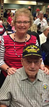 An elderly man with a black hat stating "World War I Veteran" sits at a table. His daughter stands and smiles behind him.