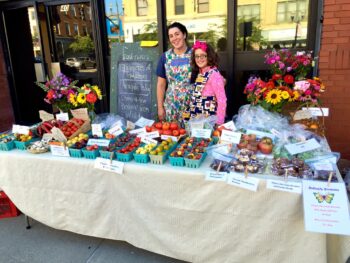 A mother and daughter in aprons stand behind a table full of fresh produce with some small bouquets of flowers in the background.