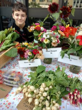 A small boy smiles next to fresh garlic and flowers.