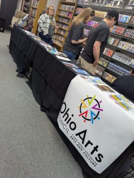 A table covered with the free comics and an Ohio Arts Council logo. Customers shop in the backgorund.