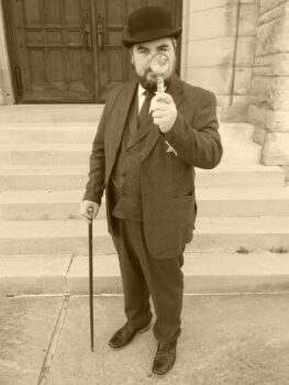 A man with a short beard holds a magnifying glass. He wears a black bowler cap and three-iece suit. He wears a badge, a pocketwatch on a chain, and holds a cane.