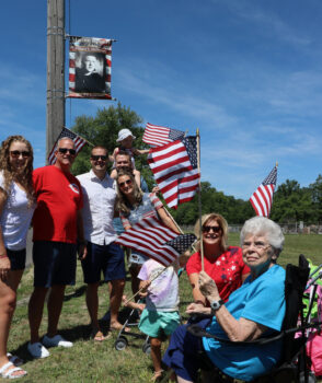 A family smiles and waves flags in front of a banner.