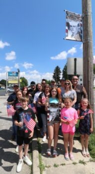 A large group stands in front of a military banner waving flags.