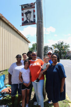 A Black family stands next to the banner of a relative and smiles. 