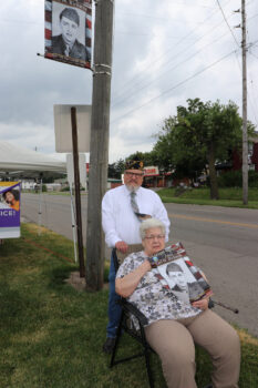 A man in a service cap stands in front of a banner. A woman accompanying him holds a banner.