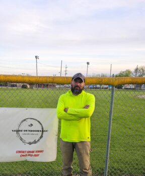 A man with a beard, hat, and a neon shirt stands next to a banner with his company's logo.