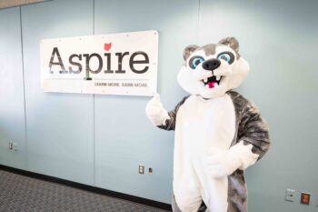 A giant gray husky mascot gives a thumbs up in front of an Aspire sign.