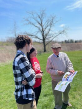 An older man points to a book while two students listen about the history of the prairie.
