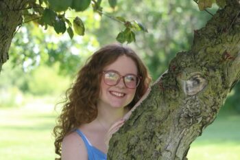 A teebage girl with long, curly red hair, glasses, and a smile stands next to a tree.