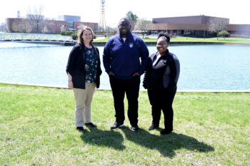 Two women and one man smile in front of the campus pond.