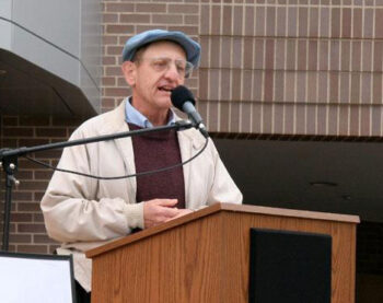 A man with a hat and glasses speaks at a podium about the prairie.