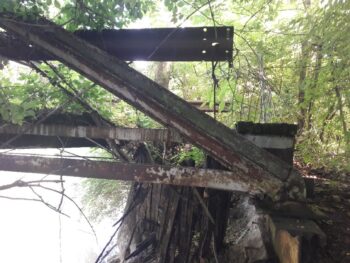 A close up shows the stseel is rusted. Some boards have rotted away. Part of the bridge is covered with tree roots.