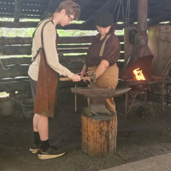 Two Scouts wearing leather aprons pound metal on an anvil. There is a fire in the background.