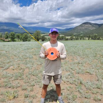 A teenage boy smiles while holding an orange circle with a long arrow stuck in it.