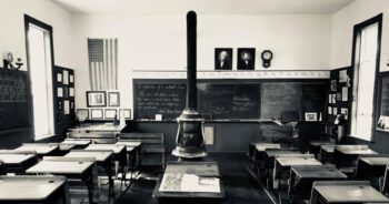 A black-and-white photo of the inside of a classroom with a stove in the middle of the room surrounded by small desks. A blackboard is behind the teacher's desk.