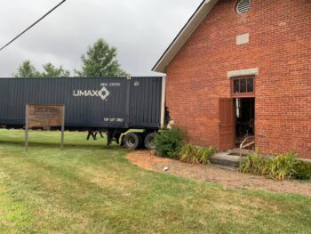 A black semi truck trailer is visible through the side of the brick schoolhouse.