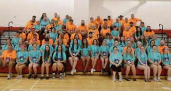 70 people in matching t-shirts smile in a gym.