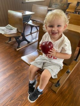 A child holds an apple and smiles while sitting in at a wooden desk. 