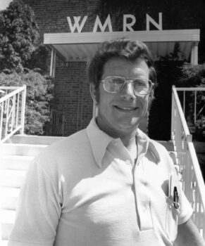 A black-and-white photo of a white man with glasses, a smile, and dark hair in front of WMRN.