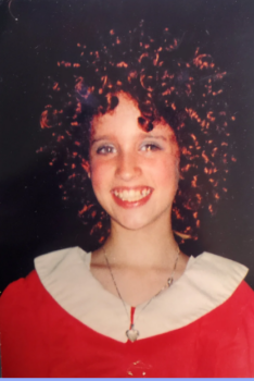 A girl with a red curly wig smiling while wearing Annie's iconic red dress with a white collar. 