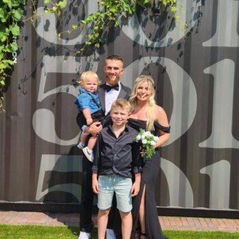 A taller man in a black suit,white button-down shirt and black bow tie; A blonde woman in a black dress floor-length dress, a blonde boy in a black shirt and shortsl and a blonde toddler in a blue denim shirt, black shorts and suspenders.