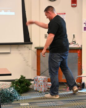 A man holds tangled Christmas lights in front of a downed tree and wrapped presents.