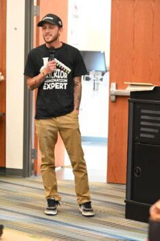Chris speaks while holding a microphone and wearing a shirt showing Christmas lights with the name "expert."