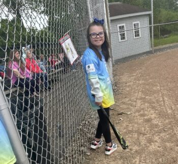 A smiling girl with long, straight brown hair; a blue pow; glasses, a jersey, holds a baseball bat inside the baseball field.