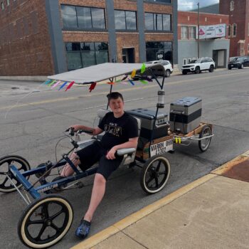 A tall boy with short hair sits on a long bicycle with two trailers hauling coolers. It has a canopy over the top. 