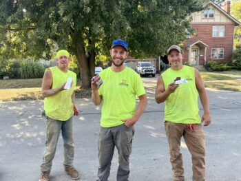 Three construction workers in bright yellow shirts smile while eating their ice cream bars.